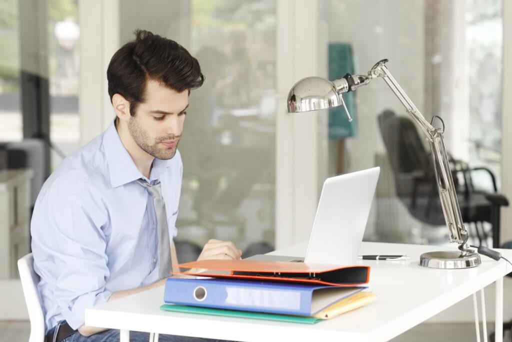 Mitarbeiter arbeitet im hellen Büro am Laptop, daneben Aktenordner